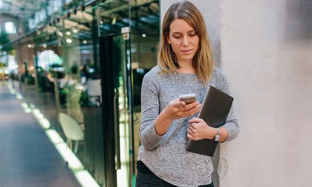 Kvinne som står i kontorkorridoren og sjekker telefonen mens hun holder en bærbar datamaskin i den andre armen