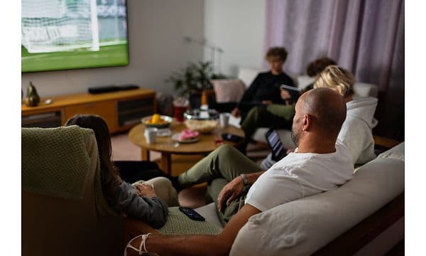 Family sitting beside a TV, some are watching TV, and some are watching their phones and iPads