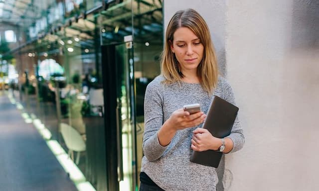 Kvinne lener seg på kontorveggen mens hun scroller sin telefonen
