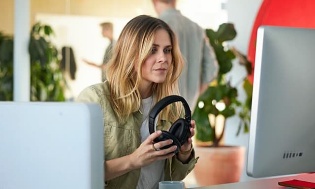 Kuulokkeita käsissään pitelevä nainen istuu tietokoneen edessä ja käyttää Microsoft 365 Office -ohjelmia työskentelyyn