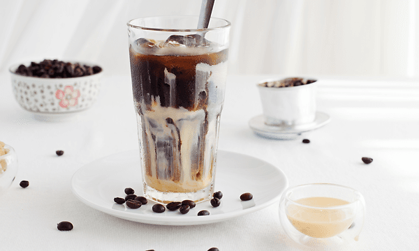A tall glass filled with ice coffee on a white table with coffee beans scattered around