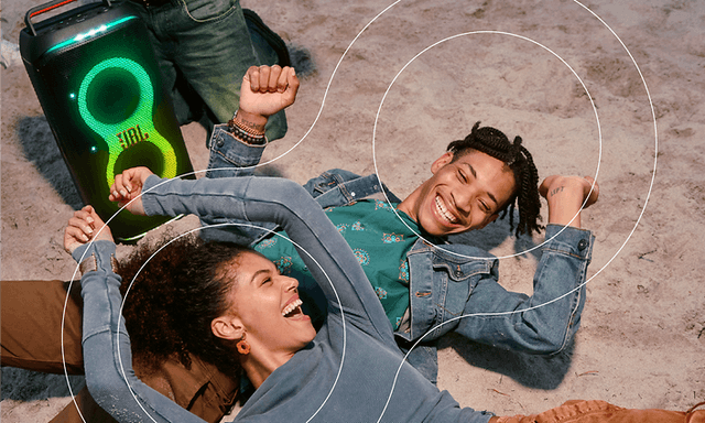 Young man and woman laying on sand next to JBL Partybox Stage 120
