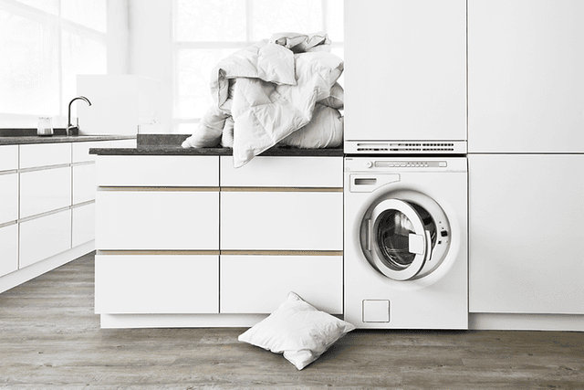 White washing room with front loaded washing machine and a pile of laundry on top of a tabletop above it