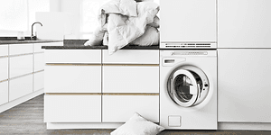 White washing room with front loaded washing machine and a pile of laundry on top of a tabletop above it