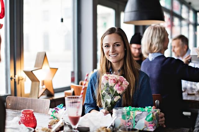 Nainen istuu kahvilan pöydän ääressä kädessään kukkakimppu ja lahjoja hänen edessään
