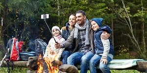 Familj som sitter på en bänk vid en lägereld och tar en selfie, flera ryggsäckar står vid sidan av. 