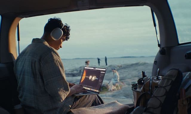 Nuori mies opiskelee rantamaisemissa MacBook Air -läppärillä Airpods Max -kuulokkeet korvissaan