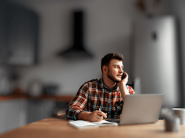 Man talking on the phone and taking notes on the paper with a laptop on the table in front of him
