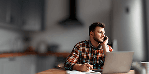 Man talking on the phone and taking notes on the paper with a laptop on the table in front of him