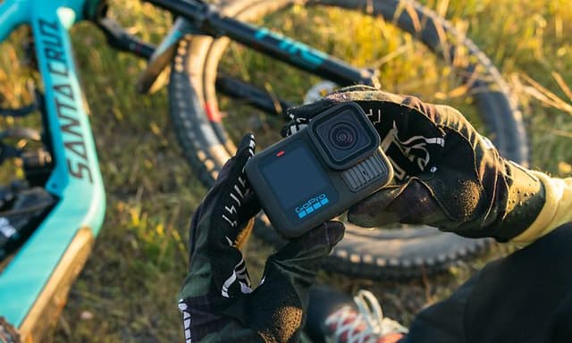 En person holder GoPro Hero 13 i hendene sine mens den står ved en terrengsykkel