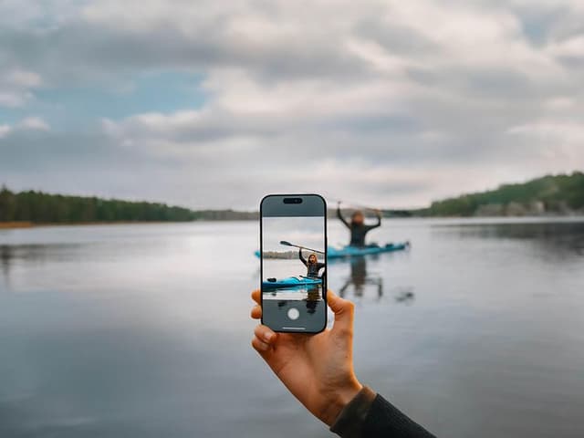 A hand using the camera control on an iPhone 16 Pro and a person in a canoe in the background