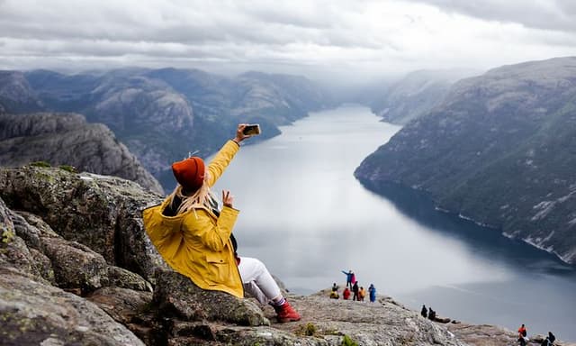 Keltaiseen takkiin pukeutunut nainen istuu kallion reunalla ottamassa selfietä puhelimellaan