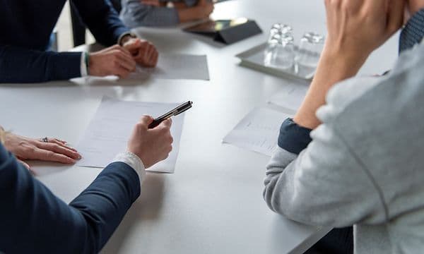 Fire personer sidder omkring et mødebord, og en af ​​dem tager noter