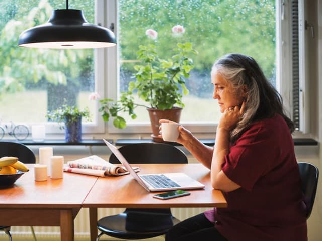 Iäkkäämpi nainen istuu käyttämässä kannettavaa tietokonetta ruokapöydän ääressä