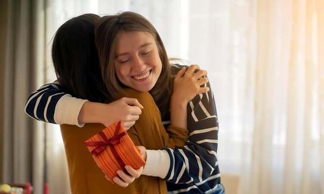 Hymyilevä nainen halaa toista naista samalla kun pitelee joululahjapakettia kädessään