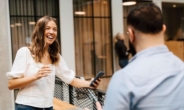Smilende kvinde, der holder en telefon, mens hun taler med en mandlig kollega