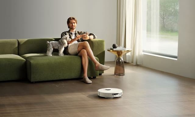 Mann og hund sitter i sofa mens robotstøvsuger rengjør gulvet