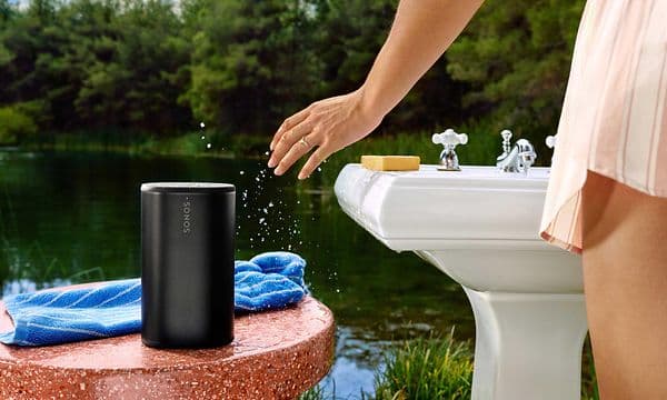 Sort Sonos Play-højtaler ved en sø, og en kvinde ved siden af en håndvask, der viser at den er vandtæt og støvtæt