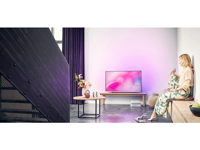 TV som sprider rosa/lila ljus i rummet. Trappa kommer ner till vänster i bild och till höger mot väggen sitter en kvinna.