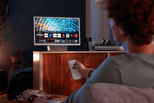 En man sitter i soffan med en vit fjärrkontroll riktad mot TV:n en bit bort, bakom TV:n döljer sig ett sovrum. 