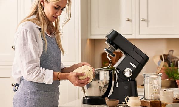 Nainen seisoo Kenwood Chef XL Pron edessä ja laittaa taikinan yleiskoneeseen