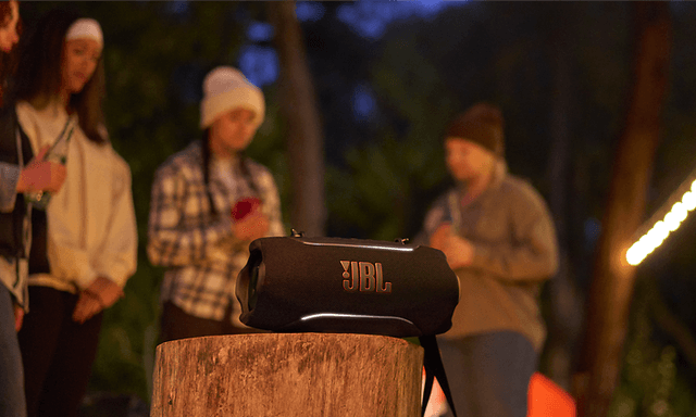 En JBL Xtreme 5-høyttaler på en trestubbe med vennegjeng i bakgrunnen.