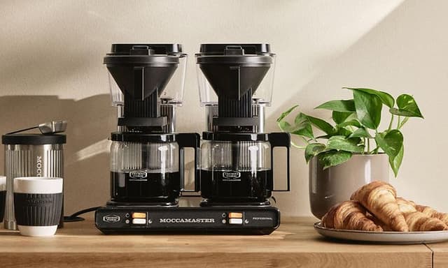 Kaksois-Moccamaster-kahvinkeitin vihreän kasvin, lautasellisen croissantteja ja valkoisia kahvimukeja vieressä