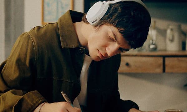 En ung man sitter vid ett skrivbord i trä, och skriver i en bok samtidigt som han bär vita Momentum 5 trådlösa hörlurar.
