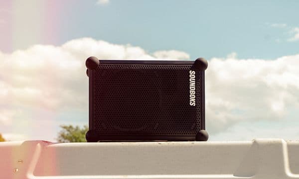 En sort Soundboks Rider-høyttaler på et hvitt gjerde, med himmel og trær i bakgrunnen.