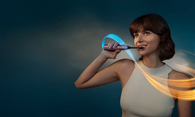 Nainen harjaa hampaitaan Philips Sonicare DiamondClean -sähköhammasharjalla sini-keltaisen valonkajastuksen ympäröimänä
