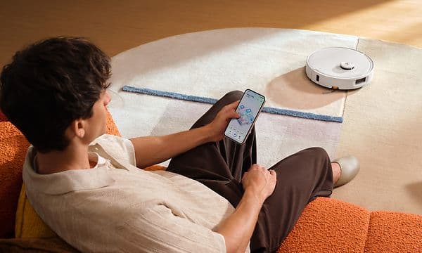 Mies rentoutuu sohvalla ja ohjaa Roborock Qrevo 2 Pro -robottipölynimuria sovelluksen kautta älypuhelimella