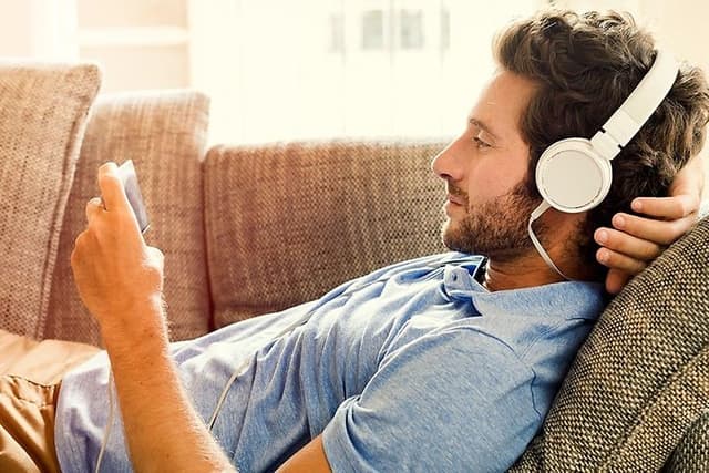 Mann ligger på sofa med smarttelefon i hånden og headset på hodet