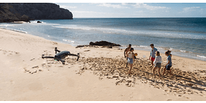 Fem personer spelar boll på en strand, till vänster synns en svart drönare som filmar händelsen ovanifrån. 