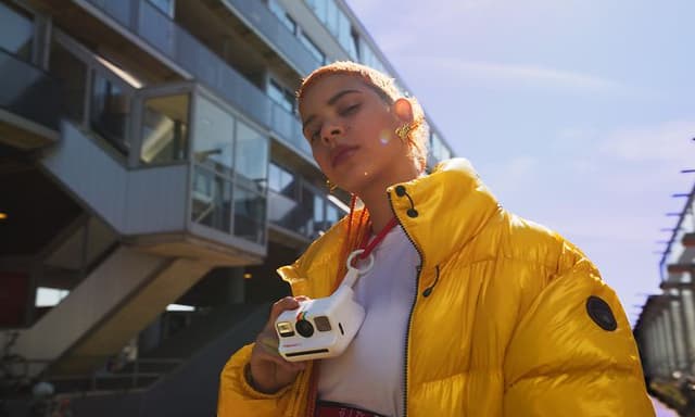 Pige med gul jakke og et Polaroid-kamera rundt om halsen.