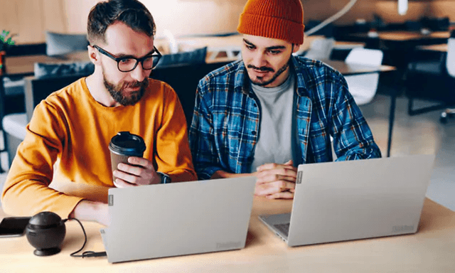 Två män sitter bredvid varandra med två bärbara datorer framför sig av modellen Lenovo Thinkbook, en håller i en takeaway kaffe.