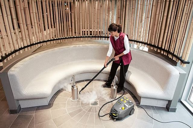 Kvinna som städar ett stengolv med en ångtvätt från Kärcher, ser ut som ett Spa eller badhus. 